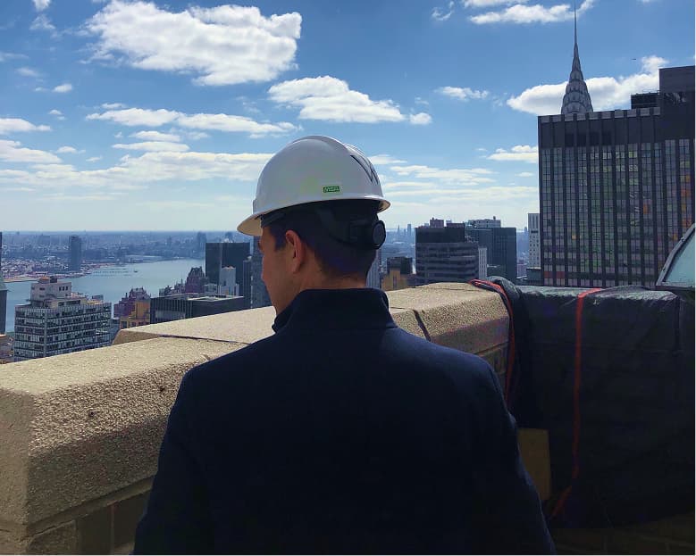 Man wearing a white construction hard hat and dark jacket standing on a rooftop terrace overlooking a city skyline with tall buildings and a river in the distance.