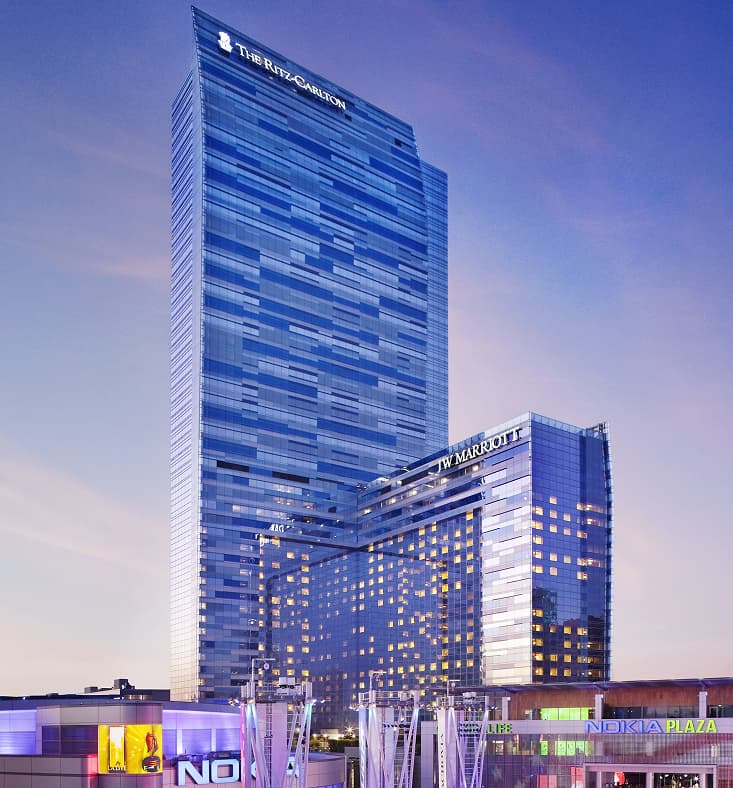Tall glass-clad high-rise buildings at dusk, including a prominent tower with ‘The Ritz-Carlton’ signage and an adjacent building with ‘JW Marriott’ signage, with lower urban development and plaza elements in the foreground.