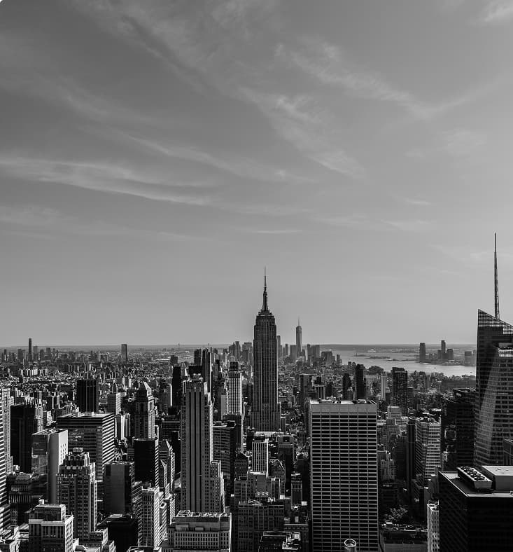 A panoramic view of the New York City skyline featuring numerous high-rise buildings including the Empire State Building near center, with a wide expanse of urban development under a partly cloudy sky.
