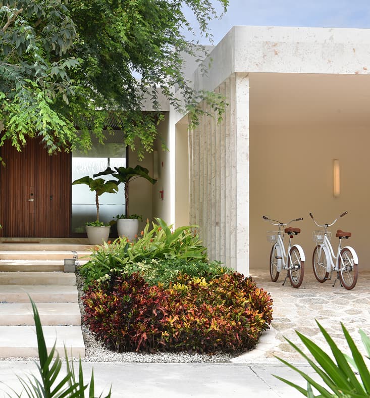 Entrance area of a luxury resort building set among lush greenery, showing a stone pathway leading to a wooden door on the left, potted tropical plants, and two white bicycles parked under a covered entryway.