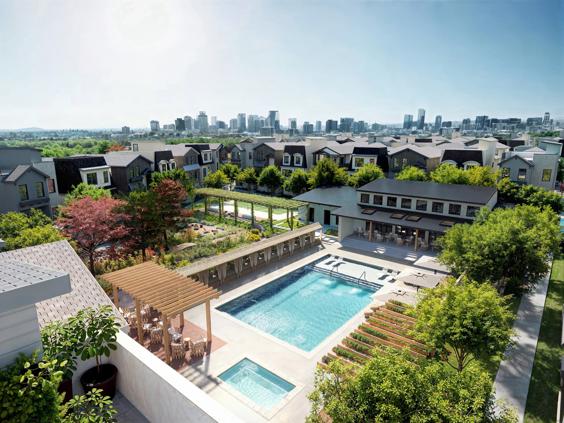 Aerial view of The Lanes residential community showing a central amenity area with a swimming pool and spa, shaded outdoor seating under wooden pergolas, landscaped gardens and trees, rows of modern single-family homes surrounding the space, and a distant city skyline under a bright sky.