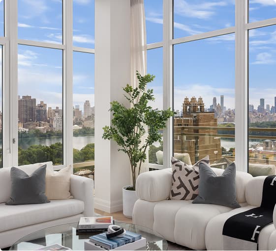 Bright modern living room with large floor-to-ceiling windows showing a city skyline and river view, white upholstered sofas with decorative pillows around a glass coffee table holding books, and a tall potted plant between the windows.