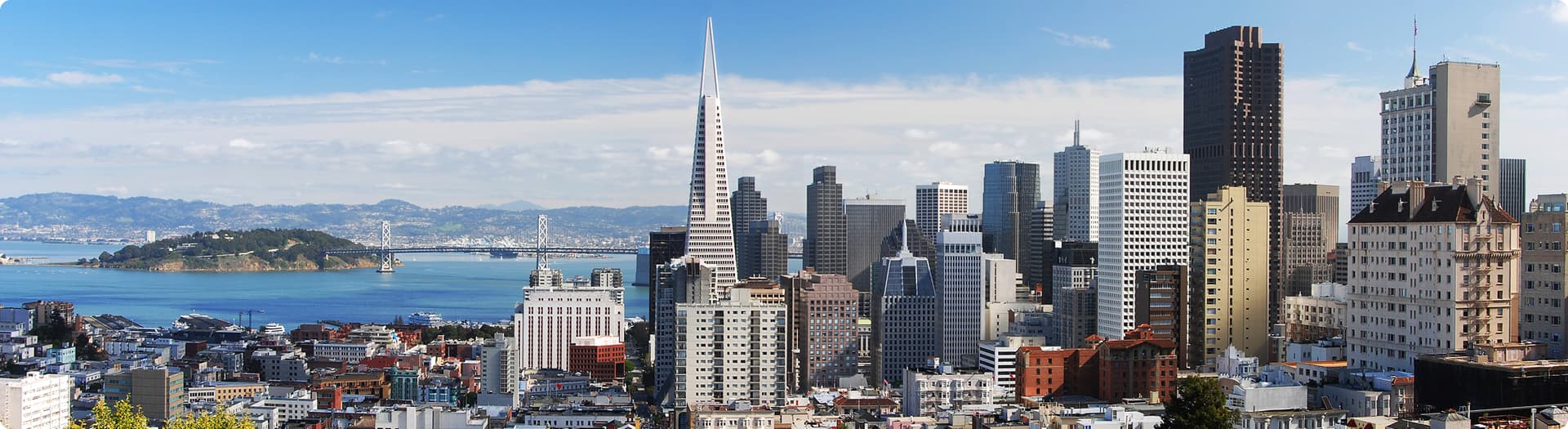 Panoramic city skyline view of San Francisco featuring high-rise buildings including the Transamerica Pyramid, with the bay and a distant bridge under a partly cloudy sky.